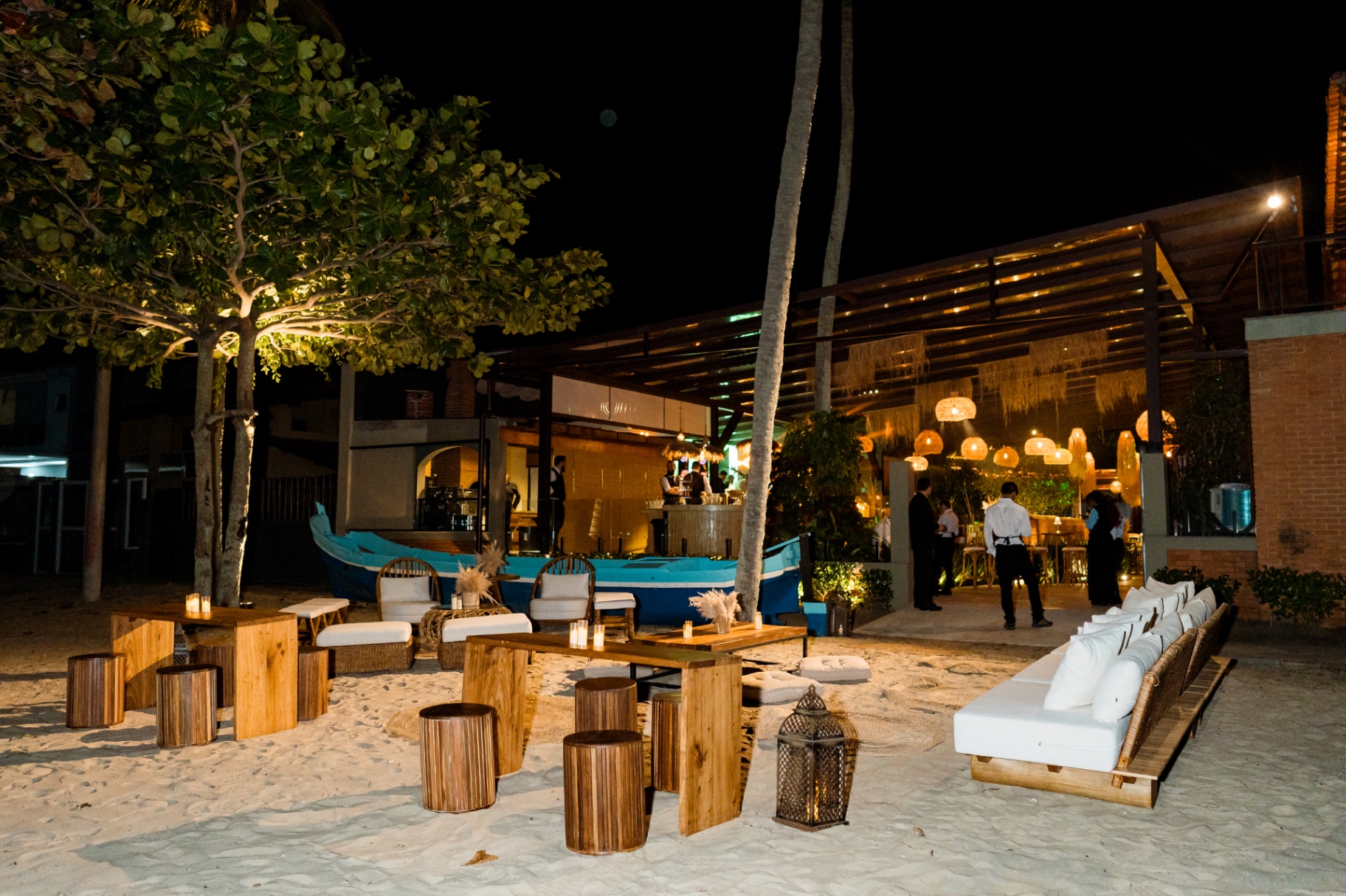 Lounge na areia da Casa Apuá com mesas de madeira, lanternas e bar iluminado à noite