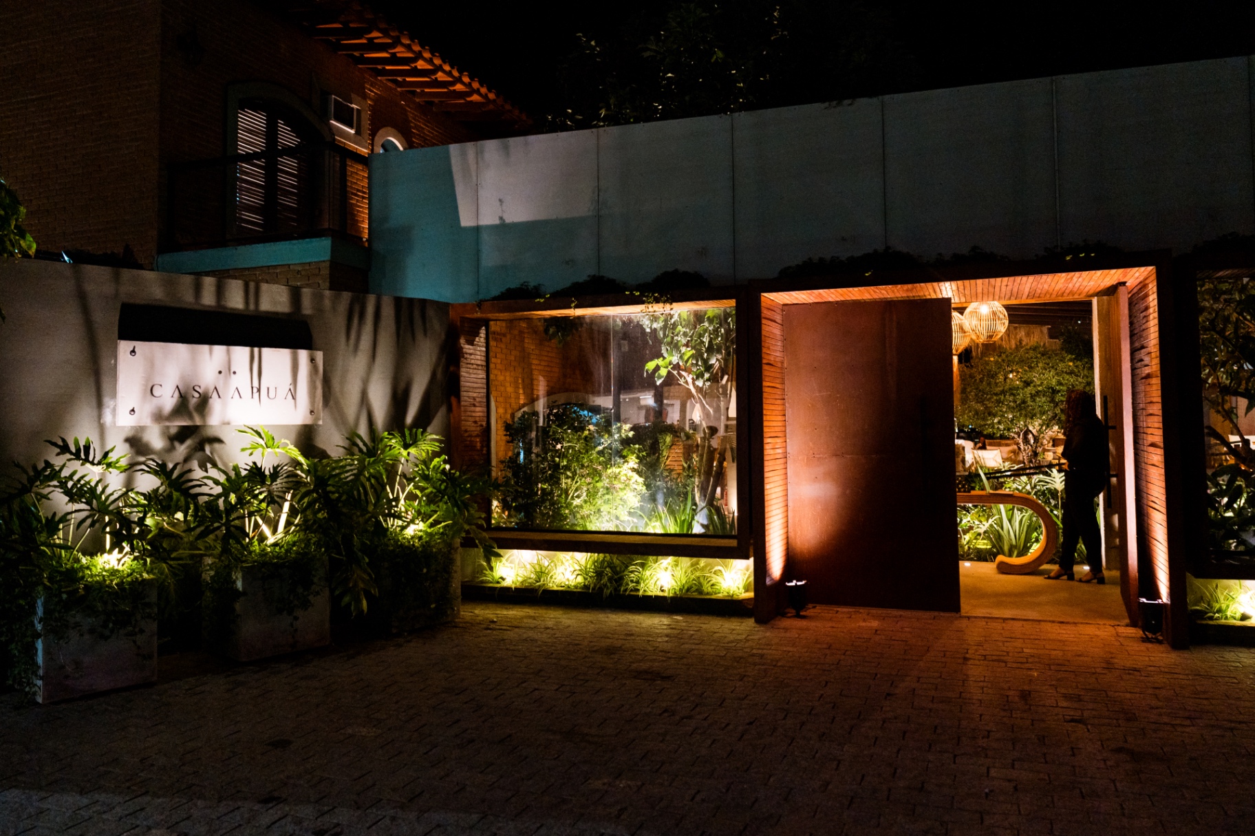 Entrada da Casa Apuá iluminada à noite, com placa do nome e jardim