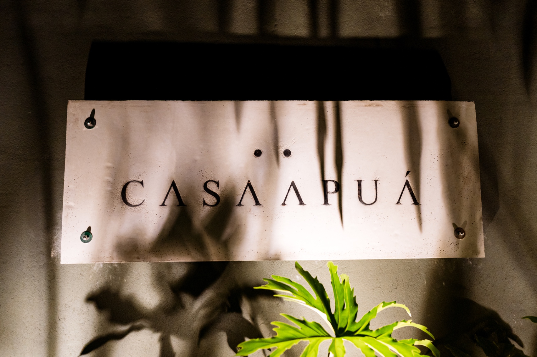 Placa de concreto com o nome Casa Apuá iluminada entre folhagens