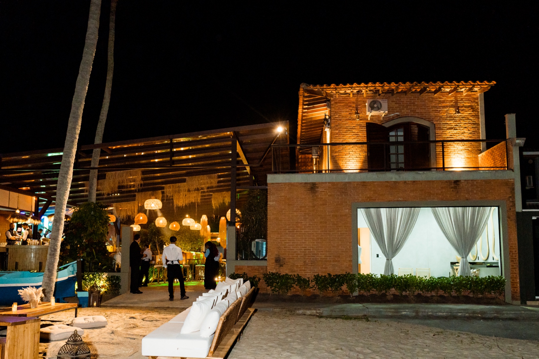 Casa Apuá iluminada à noite, com varanda de tijolo, pergolado e lounge na areia